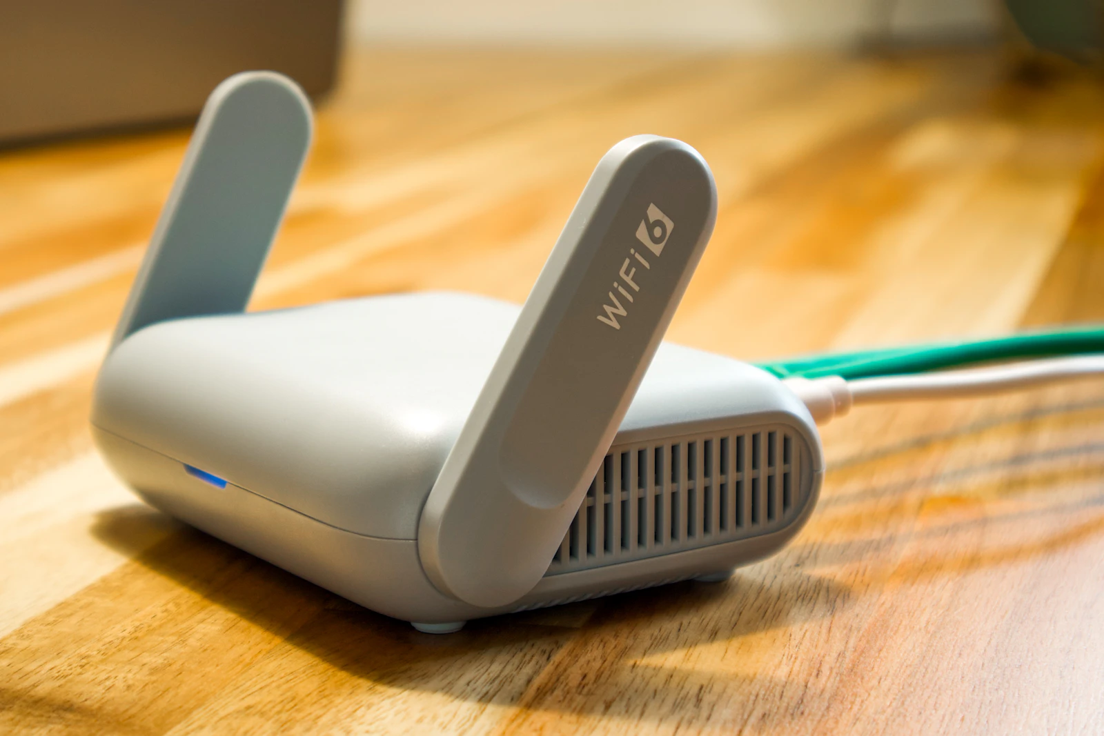 Compact grey Wi-Fi router with two antennas and a blue status light on a wooden desk, with network and power cables attached