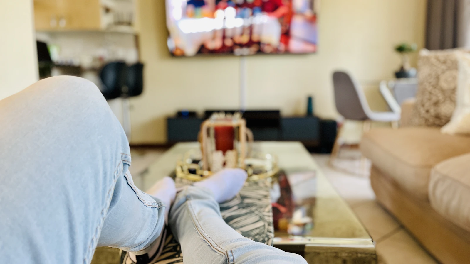 View from the sofa with feet up on a glass coffee table and a wall-mounted TV playing in a softly blurred living room